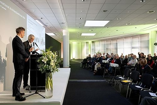 Seitliche Aufnahme einer Bühne und des Publikums. Auf der Bühne stehen zwei Männer und halten eine Präsentation. 