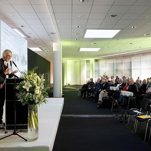 Seitliche Aufnahme einer Bühne und des Publikums. Auf der Bühne stehen zwei Männer und halten eine Präsentation. 