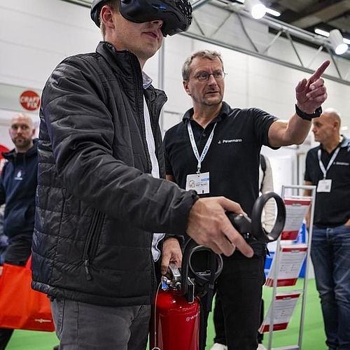 Ein Mann mit einer AR-Brille hält einen Feuerlöscher in der Hand, während ein andere Mann mit dem Finger in eine Richtung weist. 