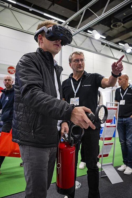 Ein Mann mit einer AR-Brille hält einen Feuerlöscher in der Hand, während ein andere Mann mit dem Finger in eine Richtung weist. 