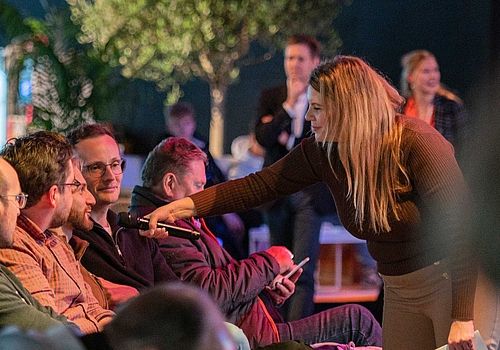 Deutsch Englisch  Situation in the GET NORD Arena: The presenter holds out the microphone to a participant in the audience so that they can actively participate in the programme.