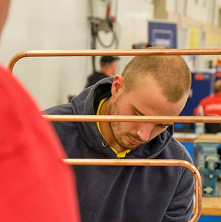 In the center of the image, a participant is working on his plumbing mastery project. You can see a bent copper pipe. Two jury members stand in front of him with their backs to the viewer.