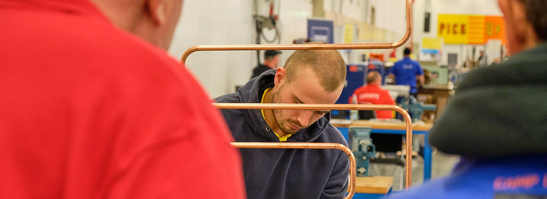 In the center of the image, a participant is working on his plumbing mastery project. You can see a bent copper pipe. Two jury members stand in front of him with their backs to the viewer.