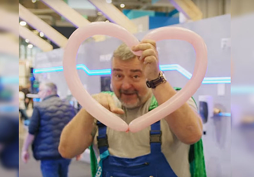 A man in blue dungarees holds up a pink balloon in the shape of a heart.