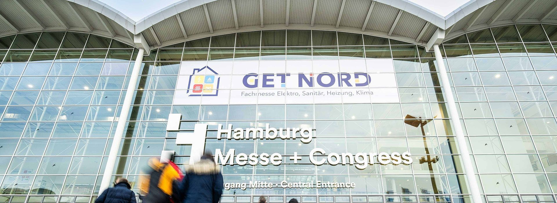 View of the facade of the exhibition building at the center entrance. A GET NORD poster is placed in the windows. In the foreground are visitors to GET NORD.