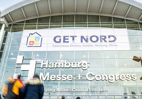 View of the facade of the exhibition building at the center entrance. A GET NORD poster is placed in the windows. In the foreground are visitors to GET NORD.