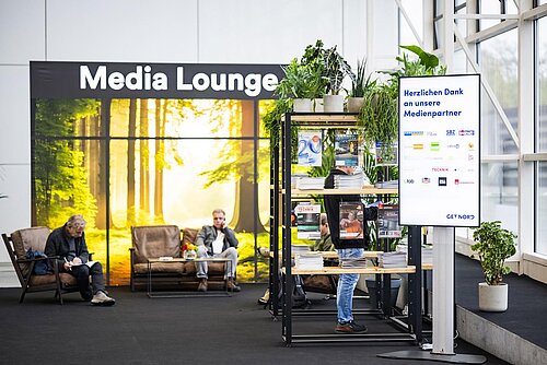 View from the center entrance to the Media Lounge. Several comfortable armchairs stand in front of a wall with a nature print and the words Media Lounge. Next to it are some shelves with magazines from media partners.