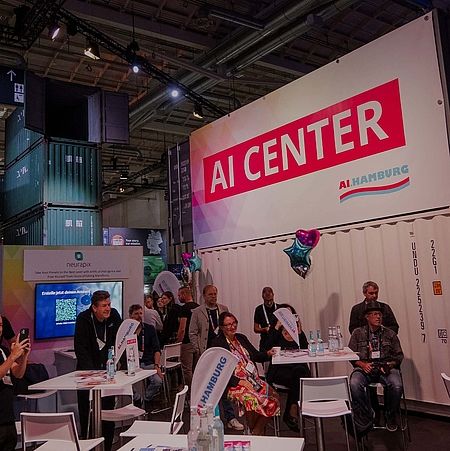 A side-view image in the AI Center Area with someone speaking into a microphone. Audience members sit at tables while one person films