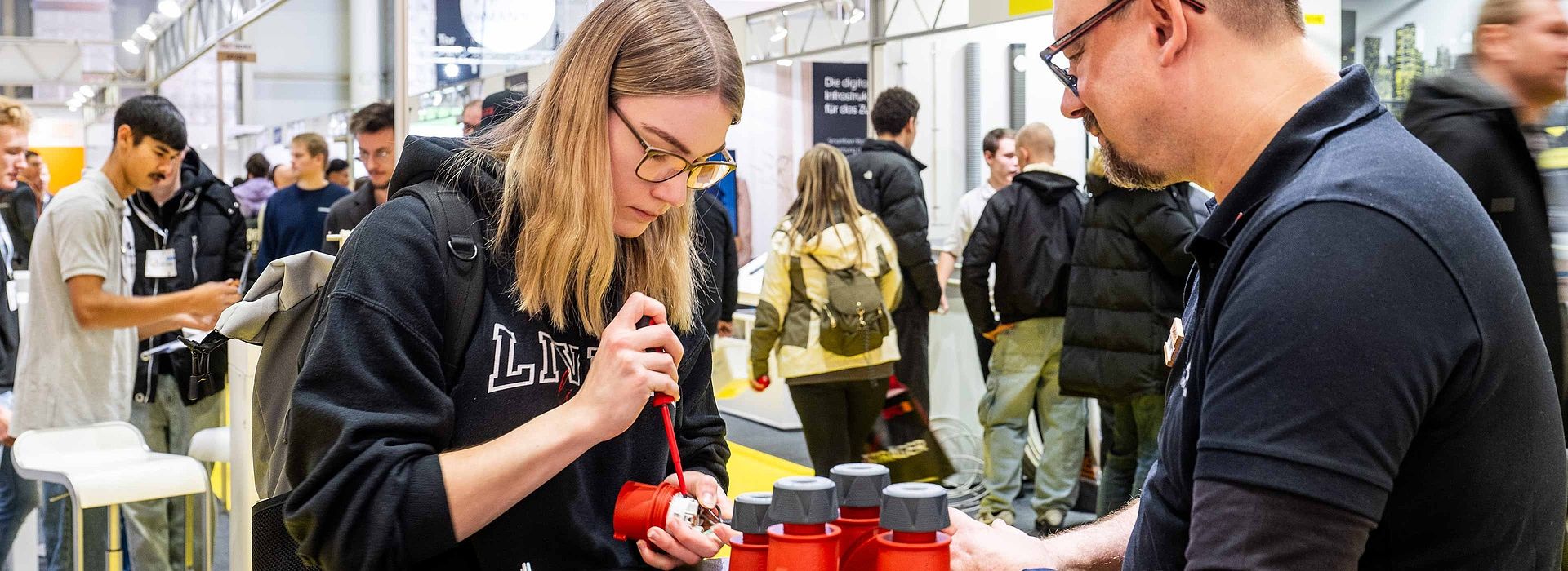 A young woman on the left side of the picture is concentrating and opening a small device with a wrench. A man on the right supports her.