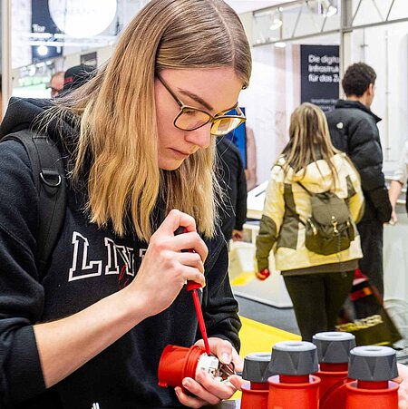 A young woman on the left side of the picture is concentrating and opening a small device with a wrench. A man on the right supports her.